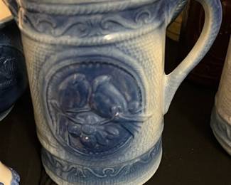 Blue Glazed Ceramic Pitcher — Large blue glazed ceramic pitcher with molded relief decoration and a generous pouring spout; a decorative and functional accent for a kitchen or dining room. The pitcher features a detailed raised pattern on the body and a comfortable handled grip, finished inside with a lighter glaze. Measures approximately for tabletop display and is sturdy but shows surface wear including small glaze chips and a couple of light scratches consistent with prior use. Good estate-sale condition overall and ready for display or functional use after a gentle cleaning.