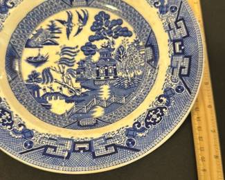 Semi China Blue Willow Decorative Plate — This Semi China decorative plate from England features the iconic Blue Willow pattern, showcasing a traditional Asian-inspired design with pagodas, trees, boats, and a bridge in vibrant blue on a cream background. The plate measures approximately 10.5 inches in diameter with a subtle raised rim and is crafted from semi-porcelain china. It exhibits light crazing and one visible chip on the rim, consistent with its age, adding to its vintage character. This collectible piece exemplifies mid-20th-century English ceramic artistry and makes a charming addition to any blue dishes collection or decorative display.