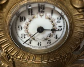 Antique Gold Cherub Mantel Clock — This charming antique mantel clock features an ornate gold-tone design with intricately detailed cherub figure and floral garland accents. The clock face has vintage black numerals and elegant minute markings set on a white background with a decorative laurel wreath. Measuring approximately 9 inches in height, 7 inches in width, and 3 inches in depth, it is perfect for display on a mantel or shelf. The clock is in fair condition with some tarnishing and wear consistent with its age, lending it added character. A beautiful decorative piece representing classic Victorian style and craftsmanship.