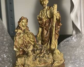 Gold-Colored Nativity Figurine — Small gold-colored nativity figurine depicting Mary, Joseph and baby Jesus on a molded base; seller notes place this in the Bonus Up Room. The piece appears to be a painted composite or resin with gilt finish and hand-painted brown highlights, showing textured ground detail and flowing robes. Approximately small tabletop size and light enough for shelf display; ideal for holiday or devotional display. Condition is good with visible paint wear and minor surface flaking to the gilt finish consistent with age and handling.