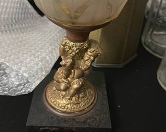 Vintage Cherub Glass Oil Lamp — This vintage oil lamp features a charming cherub base in a gold-tone finish, adding a whimsical touch to any room. The lamp has a frosted glass font with floral etching and an original glass chimney. Standing approximately 15 inches tall, it combines decorative elegance with functional design. The base exhibits some wear consistent with age, and the glass chimney shows minor dirt but no cracks. Ideal for collectors or those seeking a unique vintage decorative piece.