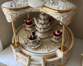 Lenox Centerpiece Teacup Ride — This Lenox Centerpiece Teacup Ride is a festive and elegant holiday collectible featuring creamy white porcelain adorned with holly motif and rich gold accents. The set includes a central teapot surrounded by four teacups, each with a Santa figure inside, all resting under a scalloped canopy detailed with embossed patterns and gold trim. Measuring approximately 11 inches in height, this centerpiece is perfect for adding a classic Christmas charm to any holiday display. It is in excellent condition with no visible chips, cracks, or wear, reflecting Lenox's superior craftsmanship. Ideal for collectors or as a unique seasonal decoration, this piece beautifully blends artistry with festive spirit.