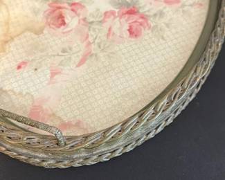 Vintage Oval Woven Tray with Roses — This vintage oval tray features a woven frame with a decorative floral insert showcasing pink roses and soft green leaves. The tray has a charming antique look with visible wear and staining on the floral fabric, adding character and vintage appeal. Measuring approximately 18 inches wide and 13 inches tall, it is perfect for decorative serving or as a display piece. The woven frame is sturdy but shows signs of age consistent with gentle use over time. A lovely piece that blends vintage charm with practical functionality for a master bedroom or living space.