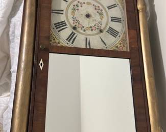 Wood & Gilt Pillar Mirror Clock — Tall wall clock with a mirrored lower door and painted dial above, featuring floral center decoration and large Roman numerals. Constructed in a wood case with paired gilt-tone turned pillars and a molded crest, the piece likely dates to the 19th or early 20th century and works as both a timepiece and decorative mirror. Overall height about 48 in with a mirrored door and visible keyhole for winding (measurements estimated from image). Condition is fair — dial shows age and surface wear, case has scuffs and the finish shows wear consistent with age; functionality unknown and pendulum/weights not shown.