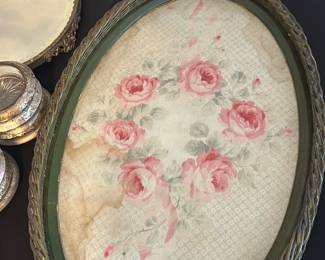 Vintage Oval Woven Tray with Roses — This vintage oval tray features a woven frame with a decorative floral insert showcasing pink roses and soft green leaves. The tray has a charming antique look with visible wear and staining on the floral fabric, adding character and vintage appeal. Measuring approximately 18 inches wide and 13 inches tall, it is perfect for decorative serving or as a display piece. The woven frame is sturdy but shows signs of age consistent with gentle use over time. A lovely piece that blends vintage charm with practical functionality for a master bedroom or living space.