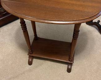 Antique Oak Oval Side Table — This antique oak oval side table features a classic design with turned legs and a lower shelf for additional storage. The warm oak finish highlights the wood grain, adding charm to any bedroom or living area. Measuring approximately 25 in high, 28 in wide, and 16 in deep, it offers a compact but useful surface. The table is in good condition with minor wear consistent with age, showcasing its vintage character. Ideal for traditional home decor and small spaces.