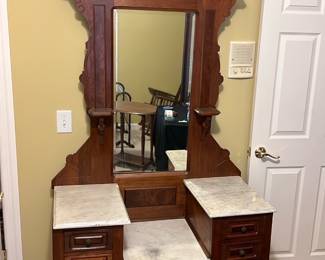 Antique Marble Top Dresser — This antique walnut dresser features intricate carved details and a large central mirror framed by ornamental woodwork. The piece includes marble tops on the side surfaces and the center vanity area, offering an elegant contrast to the rich walnut wood. Measuring approximately 68 inches tall, 42 inches wide, and 20 inches deep, it provides ample storage with multiple drawers in good vintage condition. Minor wear and patina consistent with age add to its charm without compromising functionality. This dresser is perfect for adding classic sophistication to any bedroom setting.