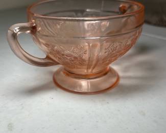 Pink Depression Glass — This vintage pink Depression glass features an elegant floral embossed pattern around the bowl and a gracefully curved handle. Made of translucent pink glass, the cup sits on a small pedestal base, typical of 1930s glassware styles. It measures approximately 2.5 inches tall and 4 inches wide including the handle, offering a charming collectible piece for any vintage glassware enthusiast. The cup is in good condition with no visible chips or cracks, though minor surface wear consistent with age is present. A lovely example of Depression era glass, suitable for display or light use.