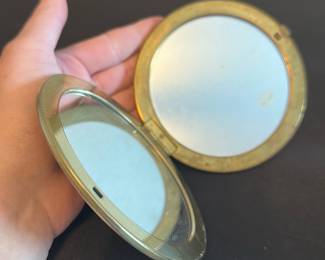 Vintage Double-Sided Round Compact Mirro — This vintage double-sided round compact mirror features a charming printed image of two girls in blue dresses with a dog, capturing a nostalgic scene on the exterior. The compact is crafted from metal with a brass tone finish, offering two functional mirrors within its hinged design. Measuring approximately 3.5 inches in diameter, it is a delicate and portable accessory suitable for personal use or display. The item shows signs of wear on the printed image but remains structurally intact and fully functional. Ideal as a collectible or a vintage beauty accessory.