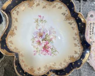 RS Prussia Blue Floral Bowl — This RS Prussia bowl features a classic blue border with intricate gold detailing and a central hand-painted floral motif in soft pinks and greens. The bowl showcases ornate scalloped edges with raised beadwork in gold, highlighting its fine craftsmanship. Measuring approximately 8 inches in diameter, this decorative porcelain piece is perfect as a display item or for elegant serving. It is in good condition with minor wear consistent with age and no visible chips or cracks. RS Prussia is renowned for quality German porcelain, making this bowl a valuable collectible from the early 20th century.