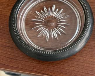 Vintage Glass & Silver Plate Coaster — This vintage round coaster features a clear glass center with a detailed starburst cut pattern and a silver-plated metal rim adorned with a beaded edge design. It serves as an elegant protective piece for tabletops, blending classic style with functional use. Measuring approximately 0.5 inches in height and 4 inches in diameter, it is compact and easily displays its intricate details. The coaster is in good condition with minor tarnish on the silver plate, typical of age and use. A stylish addition to any vintage collection or decorative serving set.