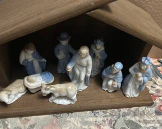 Porcelain Nativity Figurine Set — Charming porcelain nativity figurine set displayed in a simple wooden stable; figures feature soft blue and cream glazes and include Mary, Joseph, baby Jesus in manger, three wise men, shepherd and animals. The figures are small, delicately molded porcelain with glossy finish and hand-painted blue accents; stable is unfinished wood. Overall compact tabletop set good for holiday mantle or table display; figures are lightweight and easy to arrange. Condition is good overall with typical minor edge wear and a small chip on one figure base as shown.