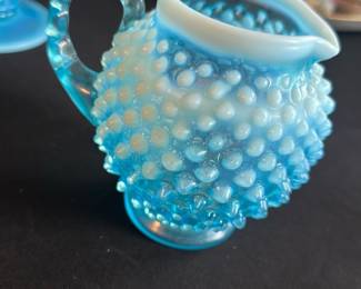 Blue Opalescent Hobnail Creamer — This charming blue opalescent hobnail creamer features a distinctive textured glass design that adds vintage farmhouse flair to any table setting. Crafted from pressed glass with a white opalescent top rim and a clear textured handle, it exudes timeless elegance and tactile appeal. Measuring approximately 4 inches tall by 5 inches wide including the handle, this creamer is the perfect size for vintage or antique dish collections. It is in excellent condition with no chips, cracks, or visible damage, preserving its classic beauty. Ideal as a collectible or functional accent piece for nostalgic glassware collectors.