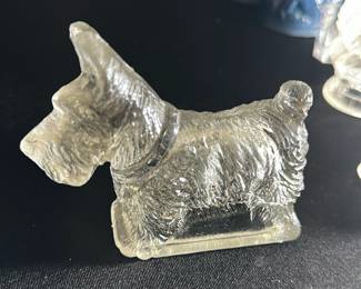 Clear Glass Scottie Dog Figurine — This charming clear glass Scottie dog figurine features detailed fur texture and a defined collar. The small collectible stands on a rectangular base, capturing the distinctive profile of the Scottish Terrier breed. Measuring approximately 2.5 inches tall, 3.5 inches wide, and 1.5 inches deep, it is a delicate decorative piece ideal for display. The figurine is in excellent condition with no chips or cracks visible. This piece adds a whimsical touch to any glass collection or dog lover's display.