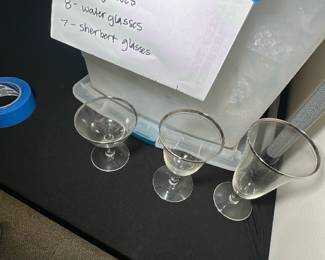 Crystal Lotus Glassware Set — This crystal Lotus glassware set includes tea glasses, water glasses, and sherbet glasses as indicated by the seller's notes. The collection features delicate stems and clear bowl shapes suitable for various drinks, making it an elegant addition to any table setting. There are 8 tea glasses, 8 water glasses, and 7 sherbet glasses stored in a labeled plastic container in the bonus up room. The glasses appear to be in good condition with no visible chips or cracks. Perfect for formal dining or special occasions.
