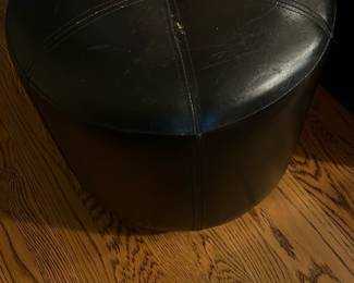 Black Leather Ottoman Stool — This black leather ottoman stool offers a versatile seating or footrest option for any living room. It features a round shape with stitched panel detail and a button-tufted center on top. Measuring approximately 18 inches in height and 16 inches in diameter, it is compact and easy to move. The condition shows visible wear and some cracks on the leather surface but remains structurally sound. Its classic design and durable leather make it a practical addition to home decor.