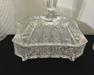 Pressed Glass Covered Candy Dish — This elegant pressed glass covered candy dish features intricate diamond and fan patterns that catch the light beautifully. The lid boasts a tall faceted handle for easy lifting, complementing the geometric design. Measuring approximately 5.5 inches high, 7.5 inches wide, and 5 inches deep, this piece adds a classic touch to any bedroom or dining room display. The dish is in good condition with no visible chips or cracks, preserving its vintage appeal. Ideal for holding candies, small treats, or trinkets, it enhances both functionality and style.