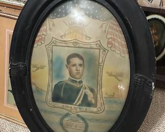 Our Hero Oval Memorial Print — Striking oval framed memorial print titled 'Our Hero' featuring a young serviceman portrait with patriotic motifs and laurel wreath. The piece has a carved black wooden oval frame (scuffs and nicks visible) with printed banners, flags and an eagle motif surrounding the central image. Measures approximately 20 in tall by 16 in wide and is a lightweight wall piece suitable for a study or gallery wall. Condition is fair with surface wear to frame, some glass glare and age-toning to the paper; a desirable patriotic memorial print for collectors of militaria and folk photography.