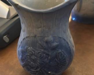 Blue Floral Pottery Pitcher — This charming blue floral pottery pitcher features an intricate embossed flower design on its exterior. The piece has a gracefully flared rim and sturdy handle, ideal for decorative or functional use in the dining room. It measures approximately 5.5 inches in height, 3.5 inches in width, and 4 inches in length, making it a compact yet elegant addition. The pitcher is in good condition, showing some wear consistent with age but no significant damage. Its classic style and glazing suggest it would complement vintage or home decor collections.