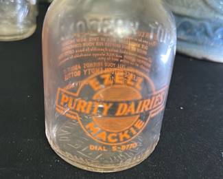 Ezell Purity Dairies Mackie Milk Bottle — This vintage Ezell Purity Dairies Mackie glass milk bottle is a charming collectible from mid-20th century dairy delivery. The clear glass bottle features bright orange vintage branding with the company name and contact dial number. Measuring approximately 6 inches in height and 3.25 inches in width, this sturdy bottle is a great display piece or addition to a dairy memorabilia collection. It shows signs of use consistent with age but remains intact without chips or cracks. A nostalgic representation of classic milk delivery, this bottle brings a touch of Americana to any collection.