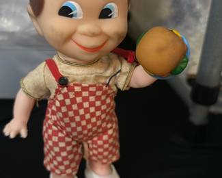 Vintage Bobs big boy burger — This vintage boy toy figurine features a charming character with brown hair, bright blue eyes, and a cheerful smile. He wears red and white checkered overalls with a cream-colored shirt and white shoes. The figure holds a round brown and blue object in one hand, adding to its playful appeal. Measuring approximately 7 inches tall, this collectible toy is in good condition with some minor wear consistent with age. A whimsical addition for collectors of vintage toys or nostalgic decor.