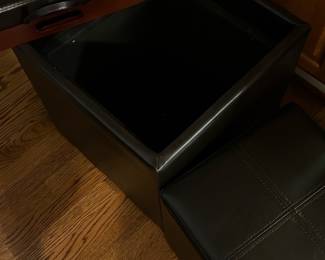 Black Faux Leather Storage Ottomans — These black faux leather storage ottomans offer a versatile combination of seating and hidden storage. Each ottoman features a hinged cushioned top with cross-stitched detailing and a spacious interior compartment for storing household items. Measuring approximately 16 inches in height, width, and length, these ottomans are lightweight and easy to move with built-in carry handles. The condition is good with some visible surface wear consistent with use but no major damage. Perfect for living room use as extra seating and convenient storage solution.