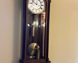 Antique Wood Wall  Clock — This antique wood wall regulator clock features a finely crafted wooden case with ornate finials and a stepped pediment design. The clock has a white face with Roman numerals and brass pendulum weights visible behind a glass door. Measuring approximately 31 inches in height, 11 inches in width, and 6 inches in depth, it is a charming addition to any dining room. The clock is in good condition with minor surface wear appropriate for its age, and the mechanical movement appears intact though not verified for working condition. This classic timepiece exemplifies 19th-century craftsmanship and makes a unique decorative as well as functional piece.