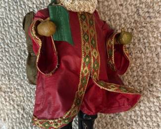 Red Cloth Santa Figurine — Decorative Santa figurine dressed in a red robe with gold-trimmed patterned ribbon and black boots. He holds a green book and wears a small sack on his back; face and hands appear painted resin or composite while clothing is fabric. Measures approximately 12 inches tall overall and is a light, collectible holiday accent suitable for mantel or shelf display. Good condition with minor wear to fabric trim and light paint loss on hands from handling.