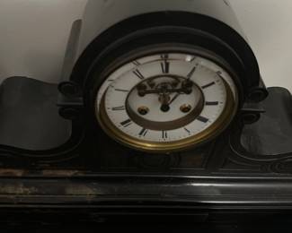 Antique Black marble Mantel Clock — This antique black mantel clock features a curved body with elegant scroll details, showcasing classic early 20th-century design. The clock face has Roman numerals and visible winding holes, framed by a brass bezel with an enamel dial. Measuring approximately 10 in high, 18 in wide, and 6 in deep, this clock is an authentic decorative timepiece ideal for a vintage mantel or shelf. It shows signs of surface wear and dust consistent with age but appears structurally sound. This piece is a fine example of traditional clockmaking, adding nostalgic charm to any bedroom or living room setting.