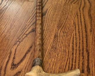 Antique Carved Wooden Cane — This antique carved wooden walking cane features a distinctive curved handle and textured shaft, showcasing fine craftsmanship. The cane is made from aged hardwood with a natural patina indicative of its vintage quality, and the handle appears to be shaped from a solid piece of antler or hardwood. Measuring approximately 36 inches in length, it is both a functional and decorative piece. The cane is in good condition with some wear consistent with age, enhancing its character. A perfect addition for collectors or those seeking a unique walking accessory.