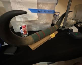 Danny Ballard 1960s Horn Display — This large decorative horn, gifted from Danny Ballard circa early 1960s at the Memphis Fair, offers a unique vintage touch for any collection or rustic decor. The piece features authentic horn with green and brown leather strap detailing, mounted on a wooden stand for display. Measuring approximately 48 inches in length with a curved and pointed shape, this horn is a striking accent piece for a bonus room or man cave. It is in good condition with natural wear consistent with its age. This collectible represents mid-20th-century Americana and is a notable conversation starter.