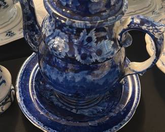 Blue Transferware Teapot and Saucer — This charming blue transferware teapot and matching saucer feature intricate floral and scenic designs characteristic of classic blue and white pottery. The teapot has a detailed curved handle, spout, and a domed lid with a finial, paired with a saucer showcasing a scenic rural estate design. Measuring approximately 6.25 inches in height and 9 inches in width including the handle and spout, this set is suitable for display or serving. The item is in good condition with some typical wear to the glaze that adds to its vintage appeal. This set represents timeless traditional ceramic craftsmanship, often found in collectible blue dishes room collections.