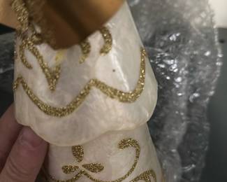 Gold Trim White shell angel — Decorative white angel figurine with gold glitter scrollwork and a metallic gold ribbon bow, ideal for holiday or year-round display. The cone-shaped layered dress appears to be a lightweight shell or resin with applied gold glitter accents and a wired halo/wing detail. Measures approximately 12 inches tall and is lightweight for easy placement; great for tabletop, mantel, or holiday vignette. In good condition with minor surface scuffs and a few small glue dots where decorative elements attach; currently wrapped in bubble wrap for storage/transport.