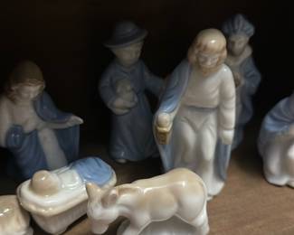 Porcelain Nativity Figurine Set — Charming porcelain nativity figurine set displayed in a simple wooden stable; figures feature soft blue and cream glazes and include Mary, Joseph, baby Jesus in manger, three wise men, shepherd and animals. The figures are small, delicately molded porcelain with glossy finish and hand-painted blue accents; stable is unfinished wood. Overall compact tabletop set good for holiday mantle or table display; figures are lightweight and easy to arrange. Condition is good overall with typical minor edge wear and a small chip on one figure base as shown.