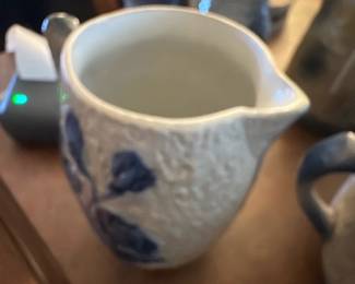 Blue Floral Ceramic Pitcher — This charming ceramic pitcher features a textured off-white base adorned with blue floral designs in relief. The piece includes a sturdy handle and a well-formed spout for easy pouring, marked with the number '18' on the base. Standing approximately 6 inches tall and 5 inches wide, this pitcher is ideal for serving beverages or as a decorative accent in your dining room. The condition is good with slight wear consistent with age but no visible chips or cracks. Its rustic farmhouse appeal makes it a lovely vintage addition to any collection or table setting.