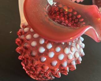 Fenton Cranberry Hobnail Pitcher — This charming Fenton cranberry hobnail glass pitcher features a distinctive ruffled rim and white hobnail detailing, typical of the Fenton Art Glass Company's craftsmanship. The pitcher showcases a clear twisted handle that contrasts beautifully with the textured cranberry body. Measuring approximately 6 inches in height and 5 inches in width, it is a perfect collectible accent for any glassware display. The pitcher is in excellent condition with no chips, cracks, or visible damage, reflecting its well-preserved quality. As a classic piece from Fenton, it represents the iconic mid-20th century American art glass tradition.