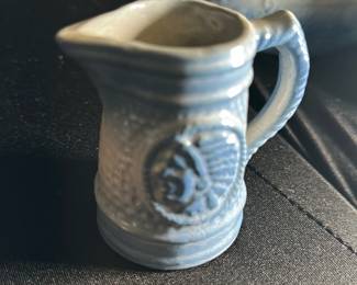 Blue Miniature Pitcher with Indian Motif — This small ceramic blue pitcher features a textured surface and an embossed Indian head motif on its side. It is crafted in a traditional style with a sturdy handle and a spout for pouring. The pitcher measures approximately 2.5 inches in height and 1.5 inches in width, making it a charming collectible or decorative accent. It is in good condition with some minor surface wear consistent with age. Ideal for collectors of miniature pottery or Americana-style ceramics.
