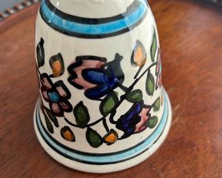 Hand-Painted Ceramic Floral Bell — This charming hand-painted ceramic bell features a colorful floral design with blue, pink, and green accents. The bell is crafted from glazed ceramic with a loop handle and a wooden clapper attached by string. Measuring approximately 4.5 inches tall and 3 inches wide, it is a decorative piece suitable for display or light use. The bell shows typical signs of age with minor surface cracks and wear but remains structurally sound. Its delicate floral motif gives it a vintage, artisanal appeal perfect for collectors or decorative use.