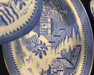 Blue Willow Covered Serving Dish — This classic Blue Willow covered serving dish features the iconic blue and white landscape and traditional Asian-inspired design. Crafted from fine porcelain, the dish includes a matching lid with a white knob accented by a gold spot, and a detailed gold rim around the edge. Measuring approximately 10 inches in diameter and 5 inches in height, it is suitable for serving or decorative use. The piece is in good condition with minor wear consistent with age and use, retaining its vibrant pattern and glaze. The maker's mark shows a crown and the letters 'SGC' stamped and embossed on the bottom, indicating quality craftsmanship.