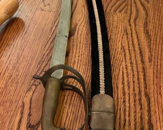 Curved Military Sword & Scabbard — Made in India. This antique curved military sword features a distinctive guard with three rings and a metal handle, complemented by a black scabbard with silver trim. The sword displays a gently curved blade typical of cavalry sabers, designed for mounted use. Measuring approximately 33 inches in length, the sword includes the scabbard which is adorned with a silver decorative stripe. The sword and scabbard show signs of age with patina and minor wear consistent with historical use but remain sturdy and collectible. This piece represents a fine example of vintage military ceremonial or functional weaponry, likely from the 19th or early 20th century.