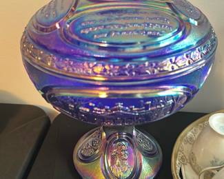 Fenton Blue Carnival Glass Eagle Covered — This striking Fenton covered dish features iridescent blue carnival glass with an eagle finial on top. The lid is embossed with inspirational text and the iconic Fenton sticker is present, confirming authenticity. The piece stands approximately 9 inches tall and 7 inches wide, making it an eye-catching collectible centerpiece. The dish is in excellent condition with vibrant coloration and no chips or cracks. A rare Fenton collectible, perfect for carnival glass enthusiasts or patriotic decor collectors.