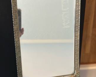 Ornate Silver-Frame Vanity Mirror — This ornate vanity mirror features an intricately detailed silver-colored metal frame with floral and scroll motifs at the top and bottom. The rectangular mirror sits within a finely embossed and patterned border that enhances its vintage charm. Measuring approximately 17 inches tall by 11 inches wide, it is perfect for a master bedroom vanity or dressing table. The mirror shows minor signs of age with some small surface imperfections but remains in good overall condition. Its elegant design is ideal for vintage or classic home decor styles.