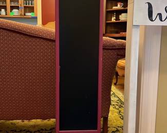 Red Standing Chalkboard Sign — This red standing chalkboard sign features a tall narrow black writing surface framed in red paint. The sign is mounted on a single pole connected to a sturdy square base, making it ideal for displaying messages indoors or in a front room setting. It measures approximately 48 inches high, 12 inches wide, and 8 inches deep, offering a slim footprint perfect for tight spaces. The item is in good condition with some minor surface wear consistent with use. Its vintage look and simple design allow it to complement a variety of home or retail decor styles.