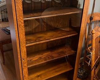Antique Oak Glass Door Bookcase — This antique oak bookcase features a glass door and an intricately carved wooden crest with an oval mirror inset. The piece offers five spacious shelves for display or storage and showcases the warm natural grain of solid oak wood. Measuring approximately 60 inches tall, 30 inches wide, and 14 inches deep, it is a perfect addition to a living room or study. The bookcase is in good condition with minor surface wear and signs of age that enhance its vintage character. Its classic design and quality construction make it a timeless furniture piece.