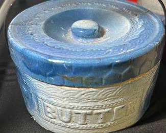 Vintage Blue Ceramic Butter Crock — This vintage blue ceramic butter crock features a textured design with the word 'BUTTER' embossed on the front. It has a metal handle with a distressed wooden grip, adding rustic charm to its classic look. Measuring approximately 4.5 inches tall and 5.5 inches wide with a 4.5-inch diameter, it is ideal for butter storage and countertop display. The crock shows some age-related wear and minor chipping around the rim but remains functional and attractive. Its sturdy construction and charming design make it a collectible piece for any kitchen or farmhouse decor.