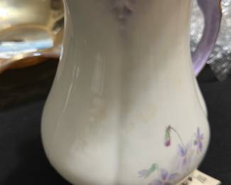 JP Vase Paris Porcelain Chocolate Pot — This delicate chocolate pot from JP Vase Paris is a beautifully crafted porcelain piece featuring elegant floral hand-painted designs in soft purples and greens. The pot has a gracefully curved form with ornate gold trim on the lid, spout, and handle, reflecting a refined vintage aesthetic. It measures approximately 7.5 inches in height, 5 inches in width, and 4.5 inches in length, making it a charming addition to any bedroom or display. The pot is in good vintage condition with minor signs of wear and no visible chips or cracks. This collectible porcelain piece bears a clear JP Vase Paris mark on the bottom, attesting to its authenticity and quality.