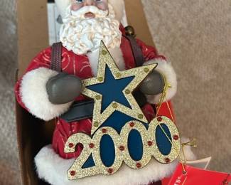 Clothique Red Santa Figure — This festive Clothiques Santa figurine from the seller notes is dressed in red with white faux fur trim and holds a glittered 2000 star ornament. Crafted from mixed materials with cloth clothing, molded face and hands, and faux fur accents, it's a display-ready holiday collectible from the Office Up Room. Overall size is compact and suitable for tabletop or mantel displays. Item appears in good condition with original tag attached and no visible damage.