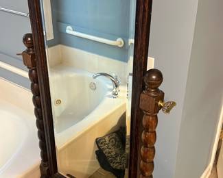 Floor Cheval Tilt Mirror — Freestanding wooden cheval tilt mirror with turned posts and decorative brass tilt keys. Dark stained wood frame with carved pedestal base and original glass, suitable for a bedroom or dressing area. Measures approximately 60 inches tall, 24 inches wide, and 16 inches deep overall; sturdy but shows surface wear and small chips on the base consistent with age. In good, usable condition aside from scuffs and finish loss at feet; brass hardware is intact and functional.