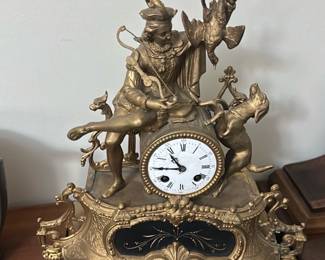 Antique Gilt Mantel Clock Figurine — This antique gilt mantel clock features an elaborate figurine of a seated hunter with dogs and a bird, showcasing intricate craftsmanship and artistic detail. The clock has a white enamel dial with Roman numerals and is set within an ornately decorated gilt metal case accented with black panels and floral motifs. Measuring approximately 16 inches in height, 14 inches in width, and 6 inches in depth, this clock is a striking decorative timepiece suitable for a traditional bedroom or living room. The clock shows some wear on the dial consistent with age, and the gilt finish has minor patina that adds character. A distinctive collectible piece typical of 19th-century French-style mantel clocks.