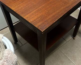 Wood End Table with Lower Shelf — Simple wooden end table with a lower shelf offers practical bedside or living-room use. Constructed in a dark stained wood with a rectangular top and straight square legs, the surface shows light scratches and wear consistent with regular use. Estimated dimensions make it a compact piece suitable for small spaces and it includes small caster feet on two legs for easy repositioning. Overall condition is fair with visible surface wear and finish loss on the top and edges but structurally sound.